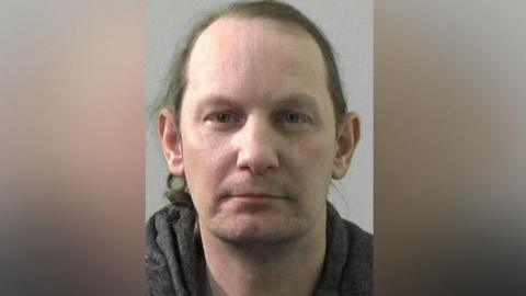 Mugshot of Winters. He is clean shaven with long straggly brown hair and wearing a dark grey hooded top.