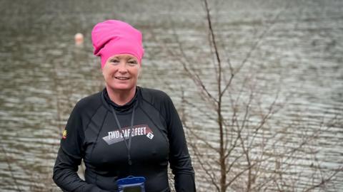 Sam Mitchell wearing a black swmming shirt and a pink swimming cap. She is standing in front of a lake