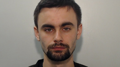 A police mugshot of Hugh Nelson. He has dark hair and a beard and has a neutral expression. He is wearing a black shirt and is standing against a plain background.
