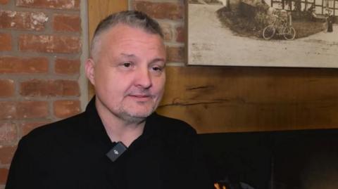A man stands in front of a fire place. He has short grey and black hair and a black shirt. Behind him is a wooden mantle piece and a red brick wall. The flames from the fire are just visible at the bottom of the image.