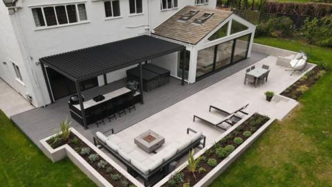 Drone shot of the patio behind the house. It is cream with a L-shaped sofa, table, pair of sun loungers, outdoor dining table and chairs. Plus a dark canopy hangs over another dining table and outdoor kitchen.