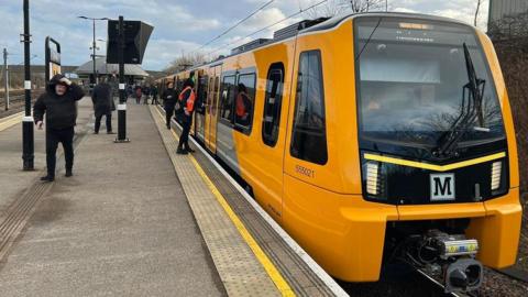 The yellow, black and grey Metro train at Pelaw station. There is a logo on the front of the train that reads: "M." And windows and doors all the way along it. People are getting on moving to alight the train. There is a dome shaped shelter further along the platform.