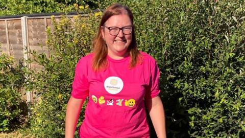 A woman with brown hair, glasses and a pink shirt standing in a garden. There is a green bush and a brown wooden fence behind her.