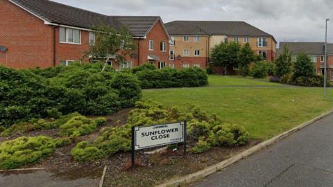 A sign saying 'Sunflower Close' on a grassy area with new-build flats in the background. 