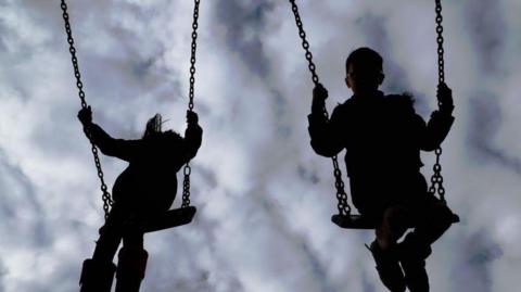 A silhouette of two children on swings against a backdrop of clouds.