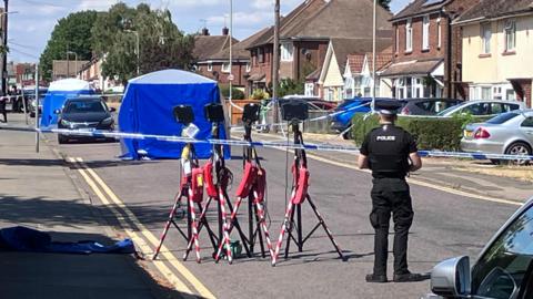 A blue police tent is set up in the middle of a residential street. Police tape blocks the road. Another tent is set up further along the road. A police officer stands with his back towards the camera as he faces the scene.