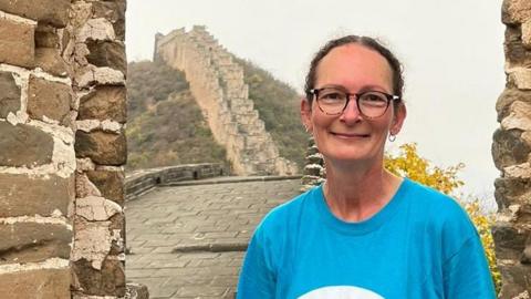 Natalie in a blue T-shirt and black trousers sitting on  wall with the great wall of China behind her