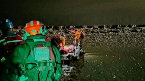 The Humber rescue team attend a casualty, who cannot be seen, on the the edge of the estuary.  The team wear helmets and hi-vis suits with "Humber Rescue" written on them. The scene is illuminated at night, revealing a muddy shore and dark water. 