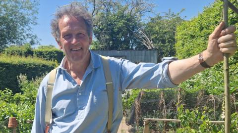 Monty Don in a blue shirt and braces holding a garden cane with one hand, in a generic promotional photo from Gardeners' World