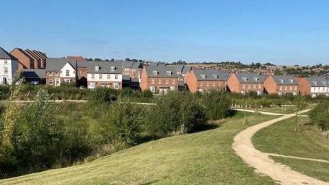A modern housing estate with grassland and shrubs in the foreground, with a path. The sky is blue.