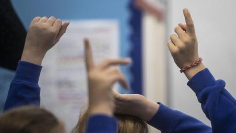 School children in a classroom with their hands up, wearing blue jumpers, foreground is blurred