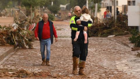 An emergency worker carries a child through a muddy street after flash-flooding