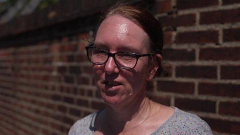 Sharon Hewitt, wearing a grey top with small blue, pink and yellow spots on, and black-rimmed glasses, looking to the left side of the camera. She has ginger hair tied back and is stood in the street in front of a red brick building.