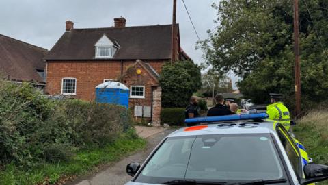 A group of police officer, mostly in hi-vis jackets, are crowded around opposite a large detached red brick house.