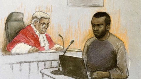 A court artist sketch of Deng Majek giving evidence with a laptop in front of him and a judge in red robes to his right