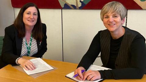 Two women sit at a desk in an office with paperwork in front of them. Julia Buckley has long dark hair and she is wearing a black jacket, pattered shirt and green lanyard. Lilian Greenwood is wearing a black waistcoat and top and holding a pen.