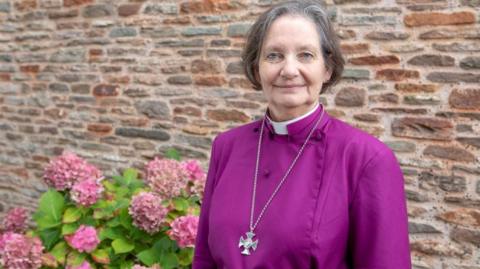 Vivienne Faull stood in front of a stone wall. She is wearing purple robes, a collar and a large silver cross around her neck. She is smiling into the camera. Pink hydrangeas are to her left.