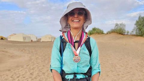 A woman in sunglasses, hat and a cyan-coloured shirt in the Sahara Desert.