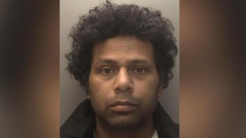 A mugshot of a man with black curly hair. He is wearing a black jacket