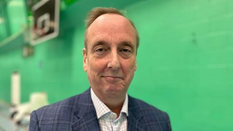 Ian Thorn smiles at the camera - he wears a blue suit jacket and light checked shirt. Blurred behind him is a green wall.