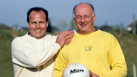 George Curtis and John Sillett: Curtis stands with his hands on Sillett's shoulders. Both smile at the camera as Sillett holds up a football