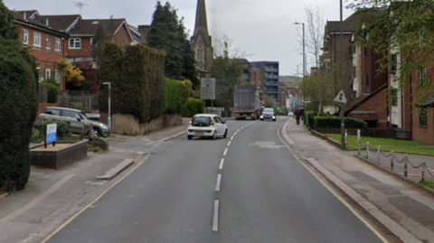 A Google Street image of Station Road in Redhill. You see a road with a small white car pulling out. There is a row of apartments on the right, a hedge on the left and a church's spire up ahead.