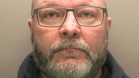 Police mugshot of a bearded man wearing dark-rimmed glasses