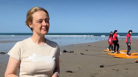 Michelle is wearing a light-coloured T-shirt stands on a sandy beach in the foreground, facing the camera. In the background, four people in red shirts practice balancing on orange surfboards laid out on the sand during a surf lesson. The beach stretches to the shoreline under a clear blue sky, with a historic stone tower visible on a small rocky islet in the distance. 