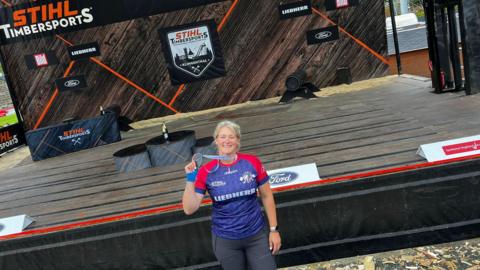 A woman with blonde hair, wearing a red and blue T-shirt, black trousers, a watch and a medal, stands in front of a wooden stage.