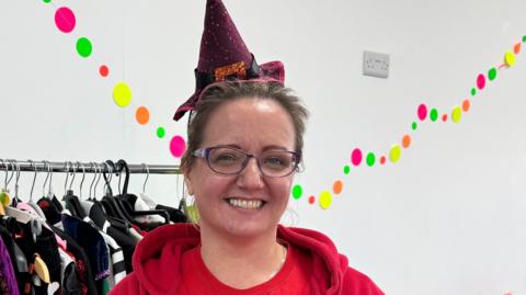 Cherry Matthews is wearing a red hoodie and a red T-shirt. She has black round glasses and brown hair. She is wearing a red spotted witch's hat with a bow on it. In the background is a rack of clothes and multi-coloured dots on the white wall. She is smiling. 