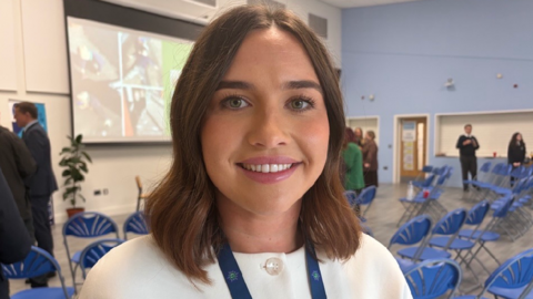 A photo of Abby Cripps. She has short brunette hair in a slight wave. She is smiling at the camera, wearing a white button up top with a blue lanyard around her neck. Behind her, she is in a school hall with blue plastic chairs and a white projector erected on the wall.