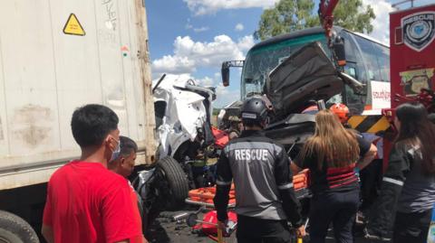 People including rescue workers wearing black helmets and black long-sleeved shirts, crowd around the scene of a vehicle accident. A bus is seen with its windshield shattered, and in front of it lies debris of crushed vehicles.