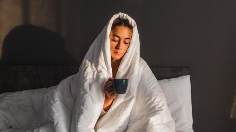A woman holds a mug in bed, with her head covered by a blanket