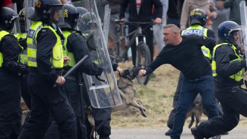 A man is bitten by a police dog during the rioting, with a line of helmet-wearing police officers holding riot shields and batons. 