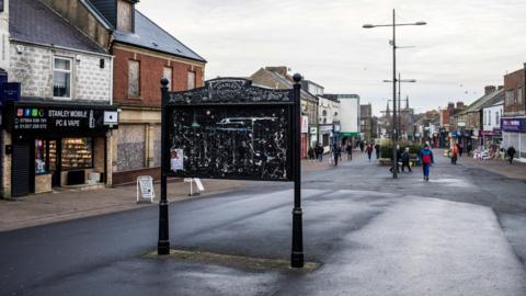 Stanley High Street. A large black notice board stands in the middle of the street. It is empty. A number of vacant and boarded up shops line the street.