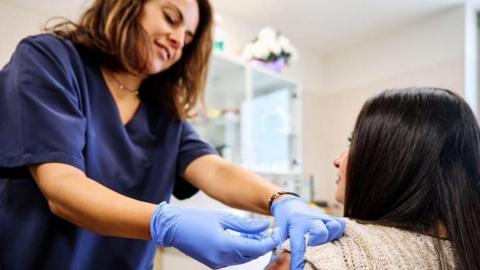 A woman dressed in a blue hospital uniform and wearing blue plastic gloves gives an injection to a woman by sticking a needle in her left upper arm. 