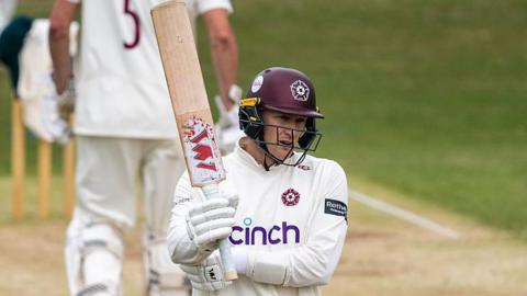 Northamptonshire batter George Bartlett, wearing white gloves, a white shirt and a maroon helmet, raises his bat