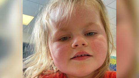 An image of a young girl looking at the camera. She is smiling and has long fair hair. She is wearing a red top.