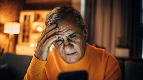 An elderly man with his hand on his head looking stressed at a smart phone
