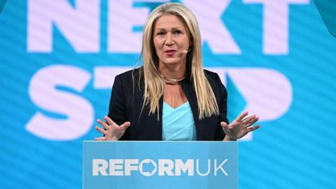 Laura Anne Jones stood in front of a screen that says Next Step in capital letters, and behind a lectern that says Reform UK. She is wearing a teal top and black jacket.