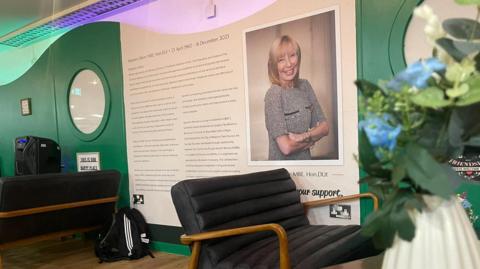 A wall of a cafe. The wall displays a photograph of a smiling woman with blonde hair cut to her shoulder. She has her arms crossed and is wearing a grey speckled dress. There is a long passage of text beside the photograph detailing her story. The rest of the wall is painted green. Padded black chairs with wooden handles sit in front of the display. Flowers can be seen in the foreground.