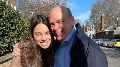 A photo of Hannah Lynch with her father Mike Lynch. Mr Lynch holds his daughter in an embrace as they smile at the camera. A road can be seen behind them with cars parked nearby and several buildings. Miss Lynch has long brown hair and is wearing a brown coat with a furred hood. Mr Lynch is wearing a navy coat with a blue jumper and shirt underneath.