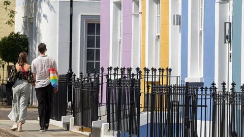 A couple photographed from behind walking down a residential street past houses painted pink, yellow and blue
