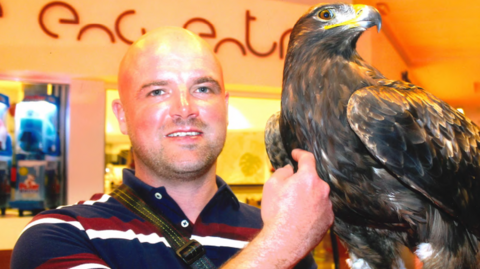 A bald man with a blue, red and white stripped shirt smiles as he strokes a large bird with his right hand.