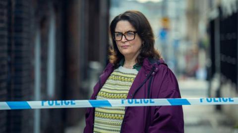 Ruth Jones as Elena Ravenscroft, in a purple jacket and glasses, stood behind white and blue police tape