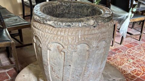 A round stone font in the shape of a barrel on a tiled floor with chairs behind it.