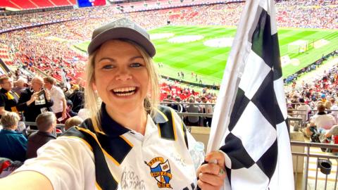 A woman holding a black and white racing flag and a football shirt stands in the stands of a large stadium. A football pitch can be seen behind her.