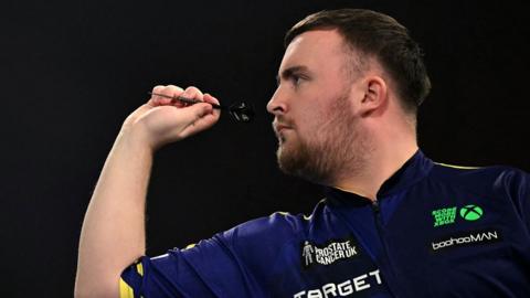 Luke Littler, a teenager with short dark hair and a light beard, faces to the left and aims with a dart 