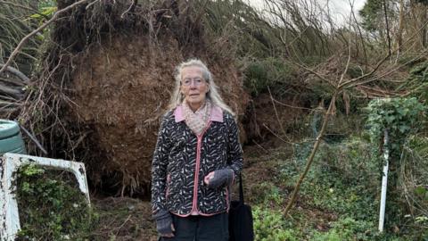 A woman stood in front of a large tree which has been uprooted and fallen over. She is wearing a black and pink fleece and is holding a bag in her left hand.