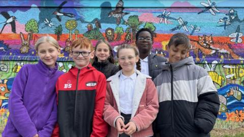 A group of six children all wearing coats and smiling for the camera. Behind you can see a colourful mural with birds, dragonflies, rabbits, bees and fish drawn on it in a wildlife setting of trees, grass and a river.